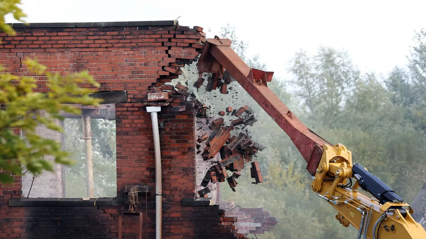 Excavator boom pushing over a brick wall of an old mill building
