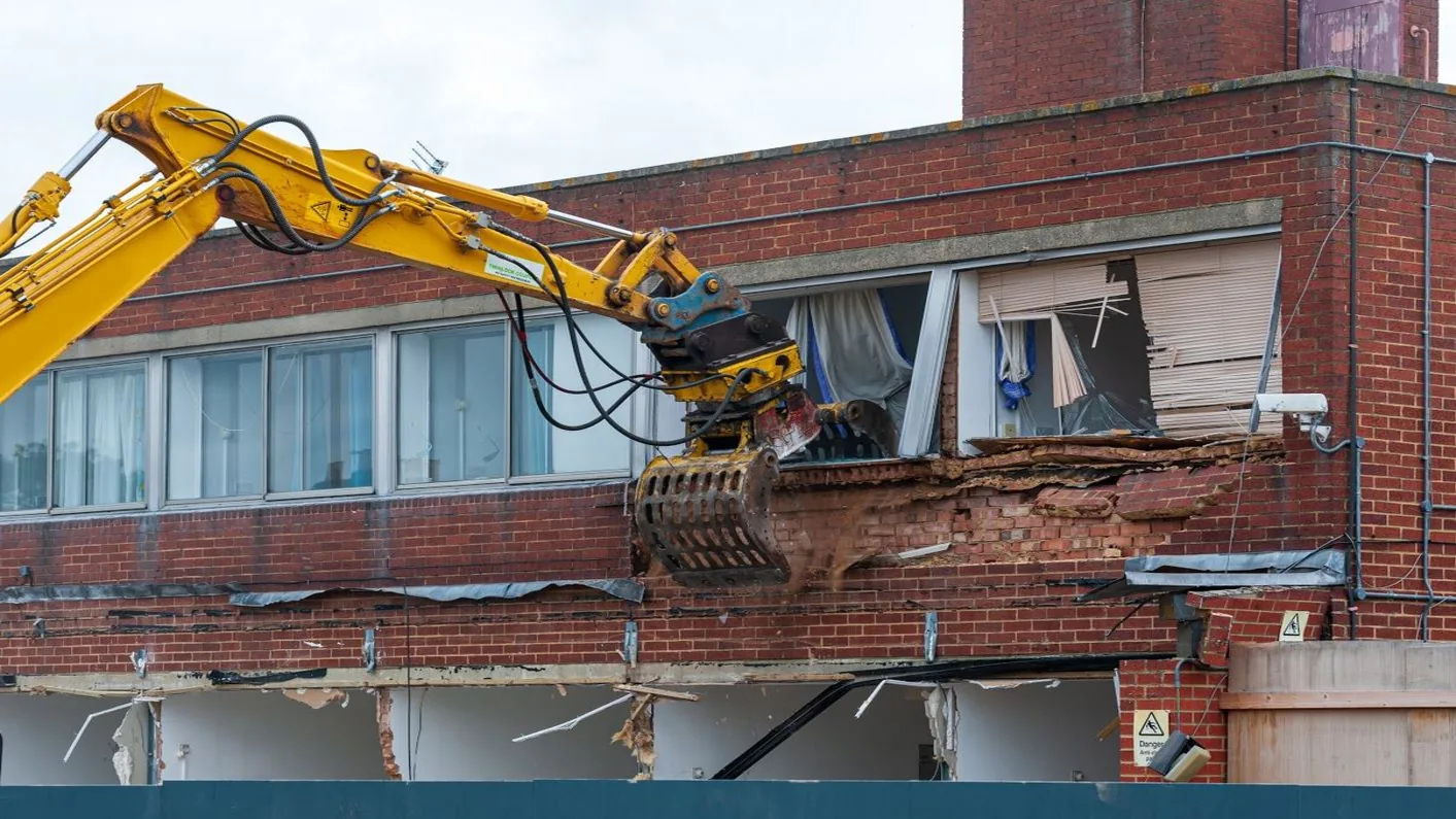 Excavator with a pulverizer attachment cutting into the brick face of a two-story building