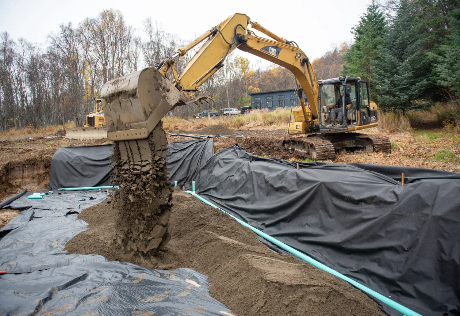 Excavator placing fill over drainage pipe and membrane along a trench