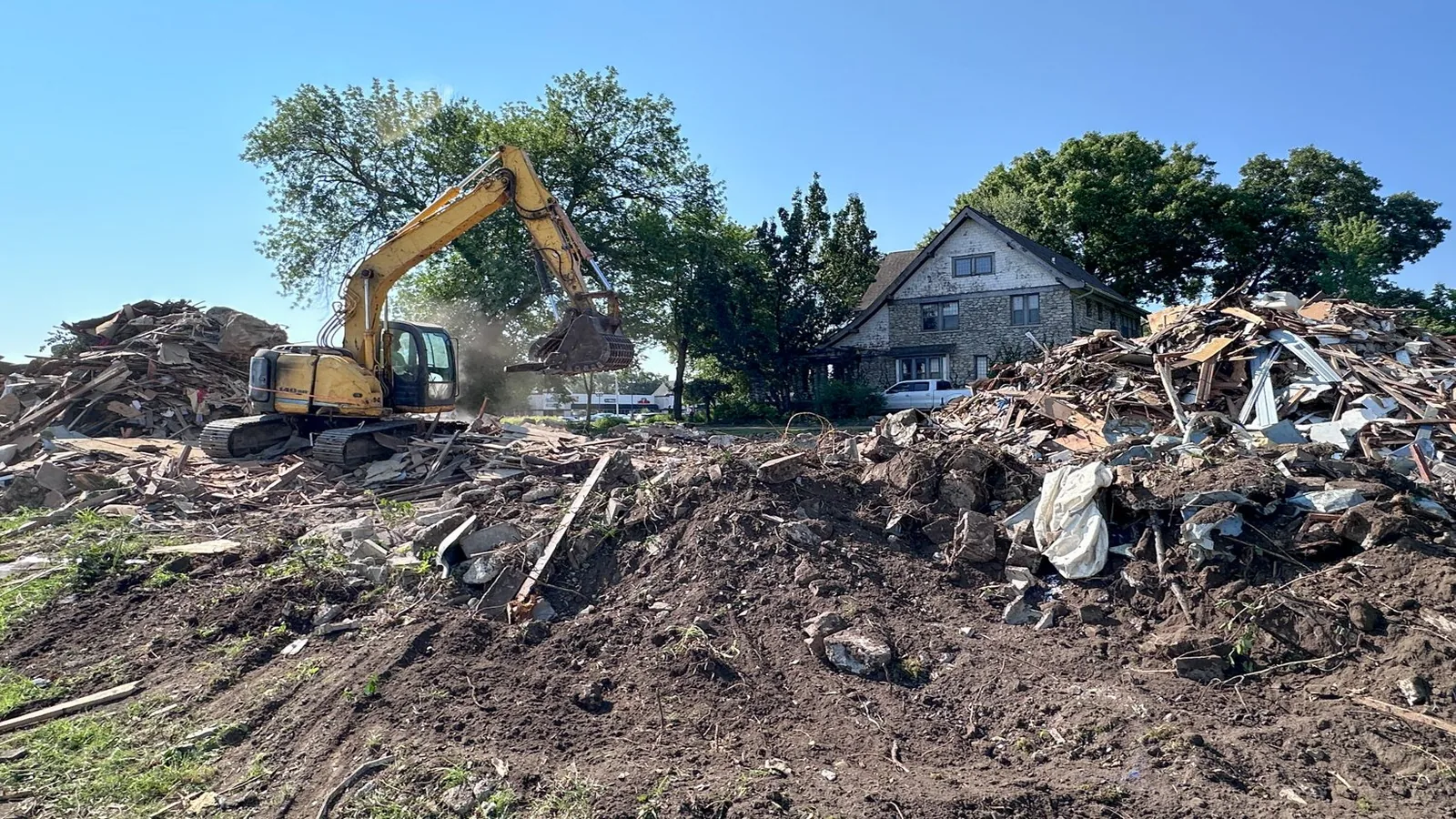 Excavator working a large demolition debris pile with a stone house behind it