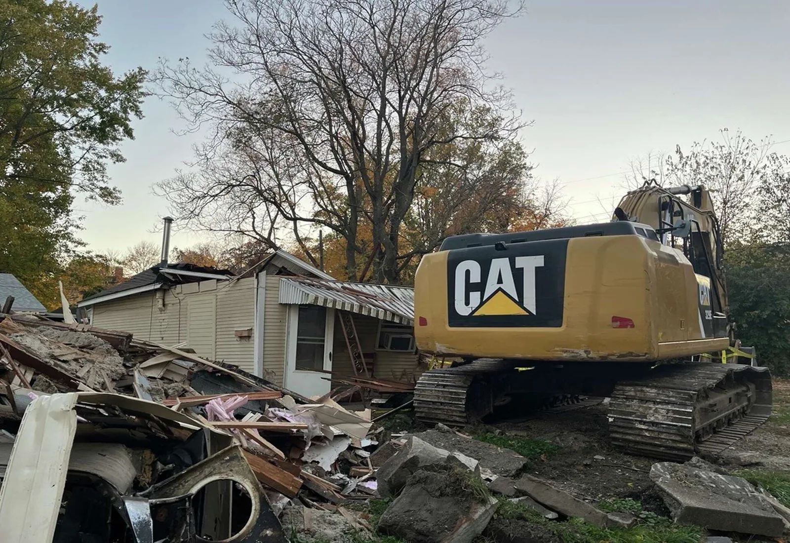 Excavator taking down a small white outbuilding with fall hardwoods behind it