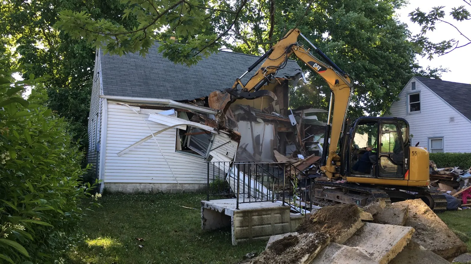 Tracked excavator tearing into the side of a two-story wood-frame house with debris on the lawn