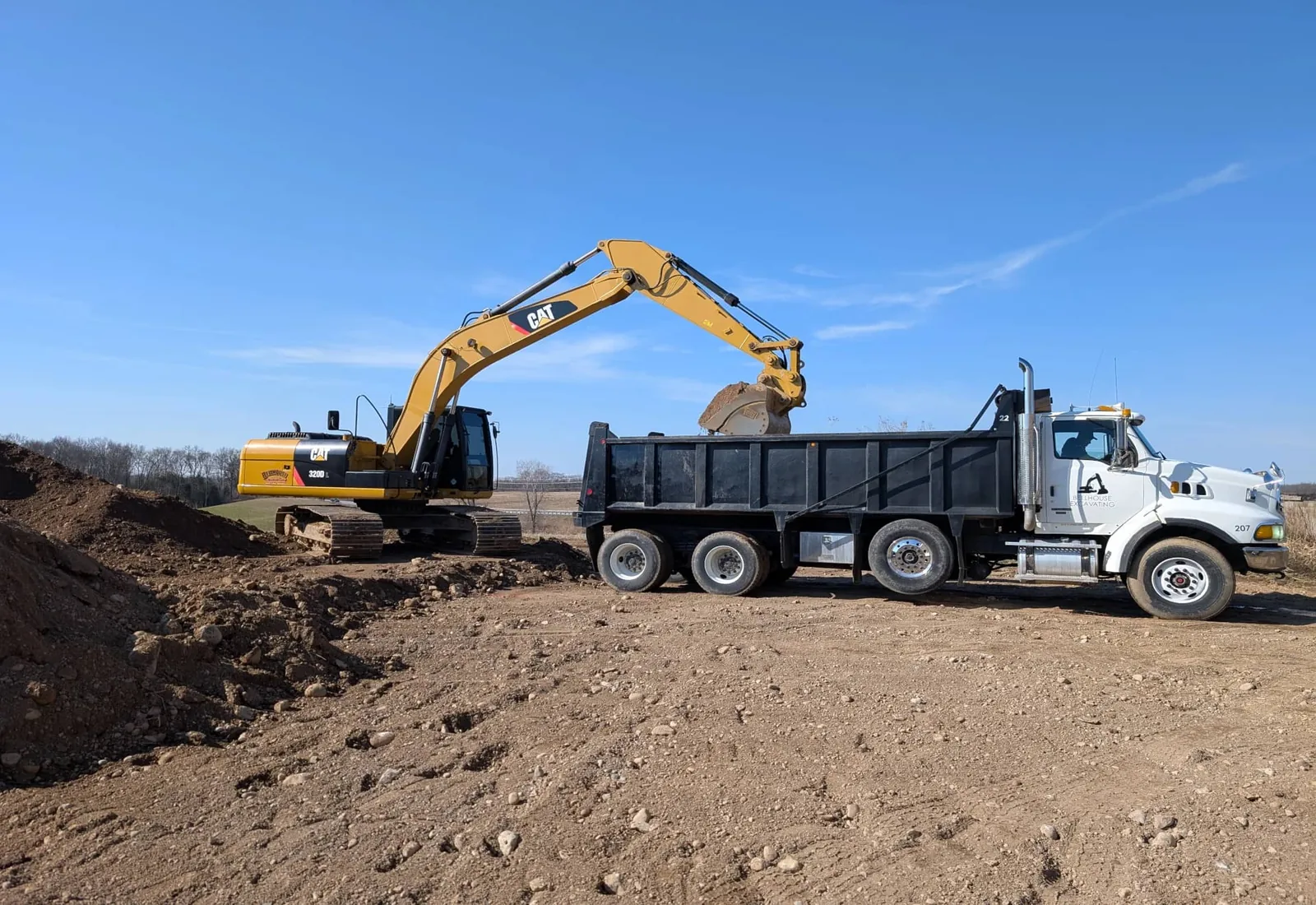 Excavator loading spoil into a tandem dump truck on a cleared lot