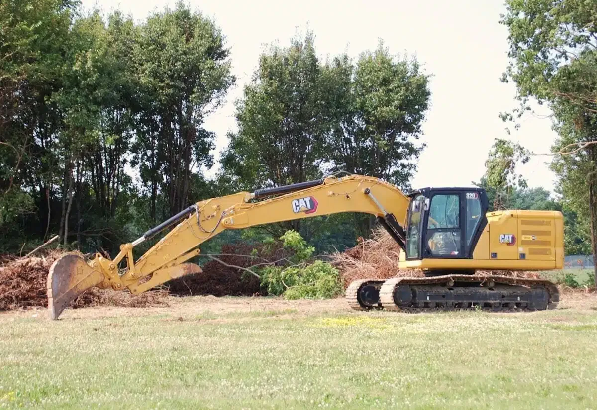 Excavator working a cleared field with brush piles and a standing hardwood tree line behind