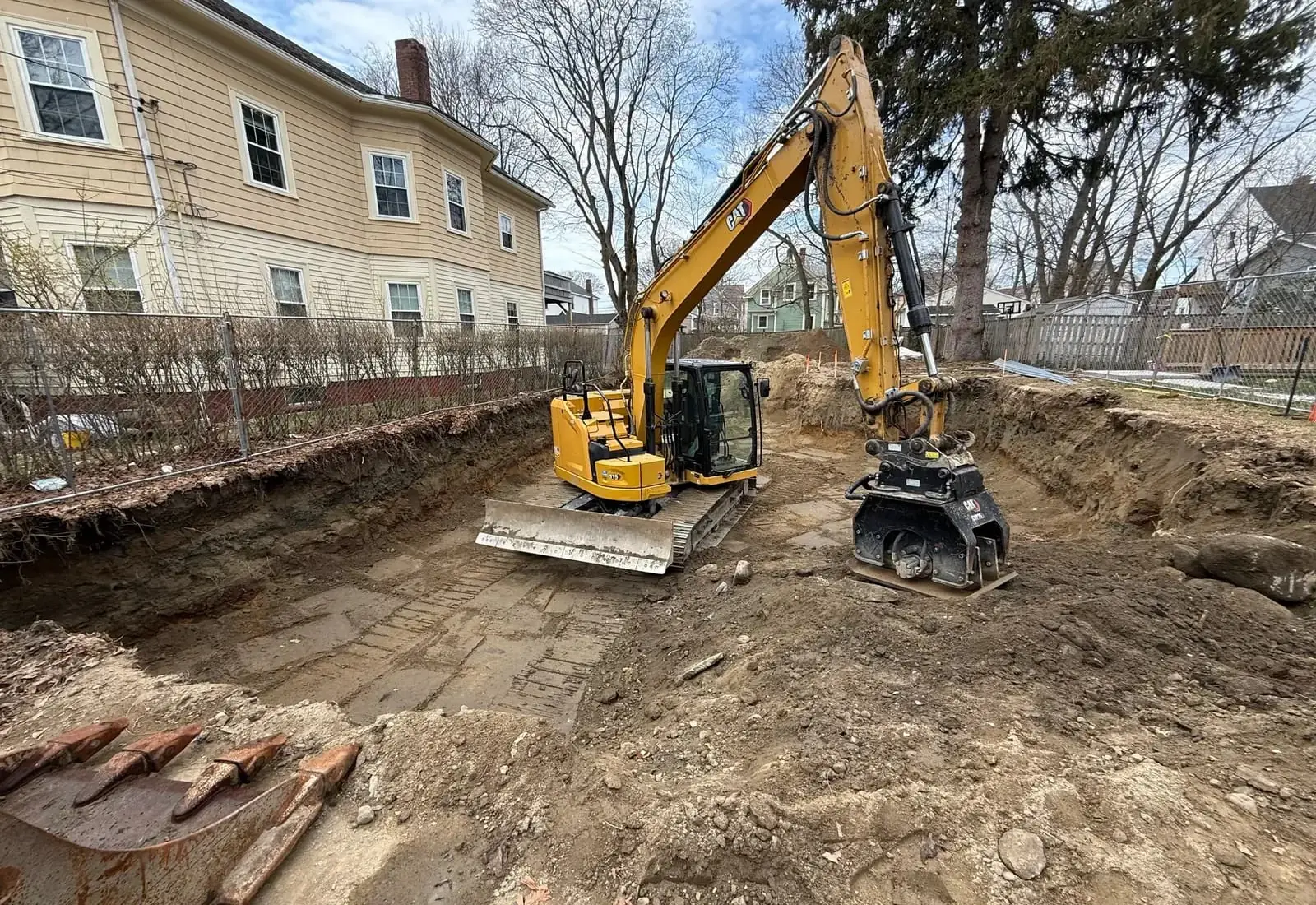 Compact excavator and a plate compactor inside a dug foundation beside a clapboard house