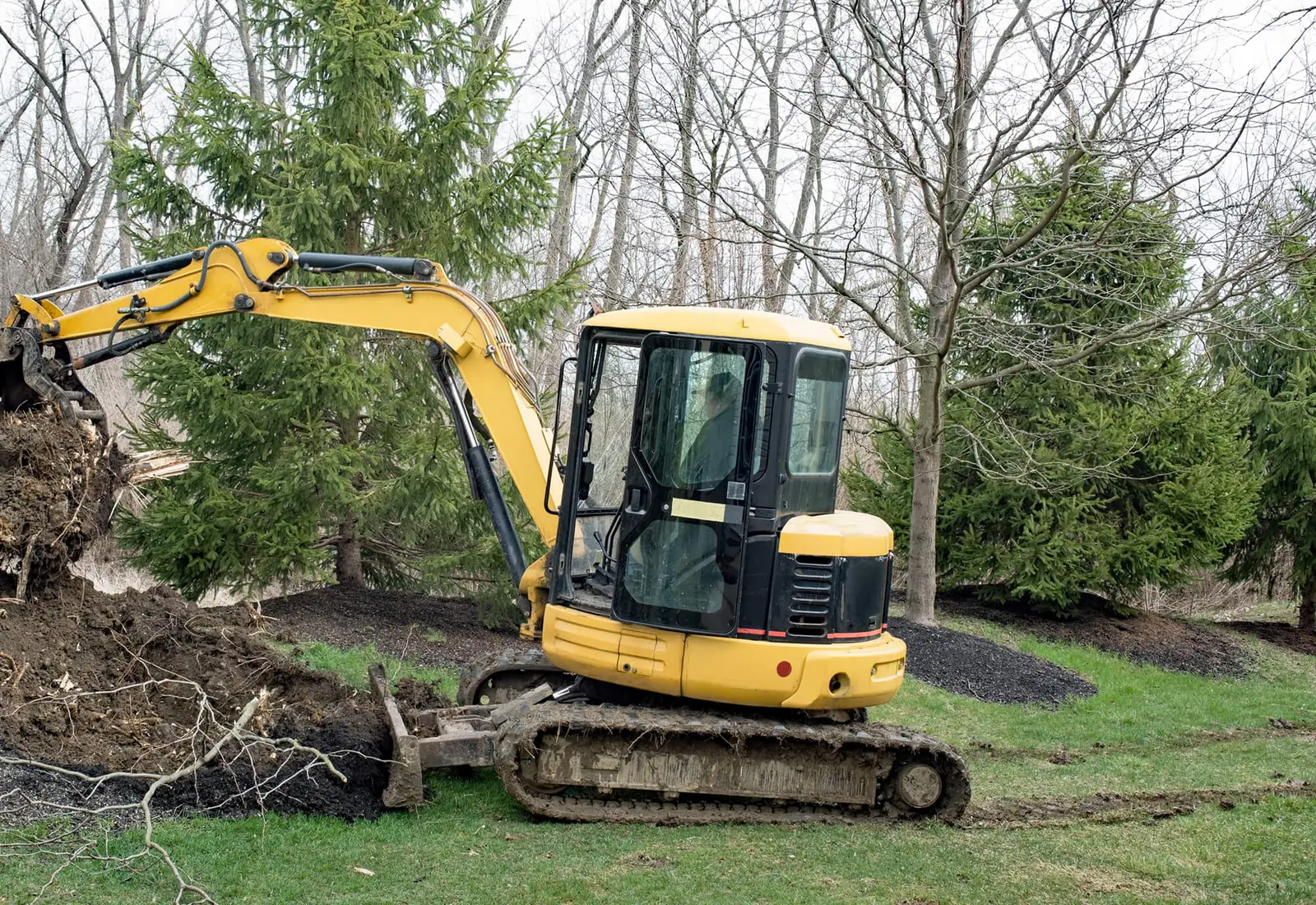 Compact excavator on a lawn working the bare dirt where a tree stump was dug out, with the root mass beside it