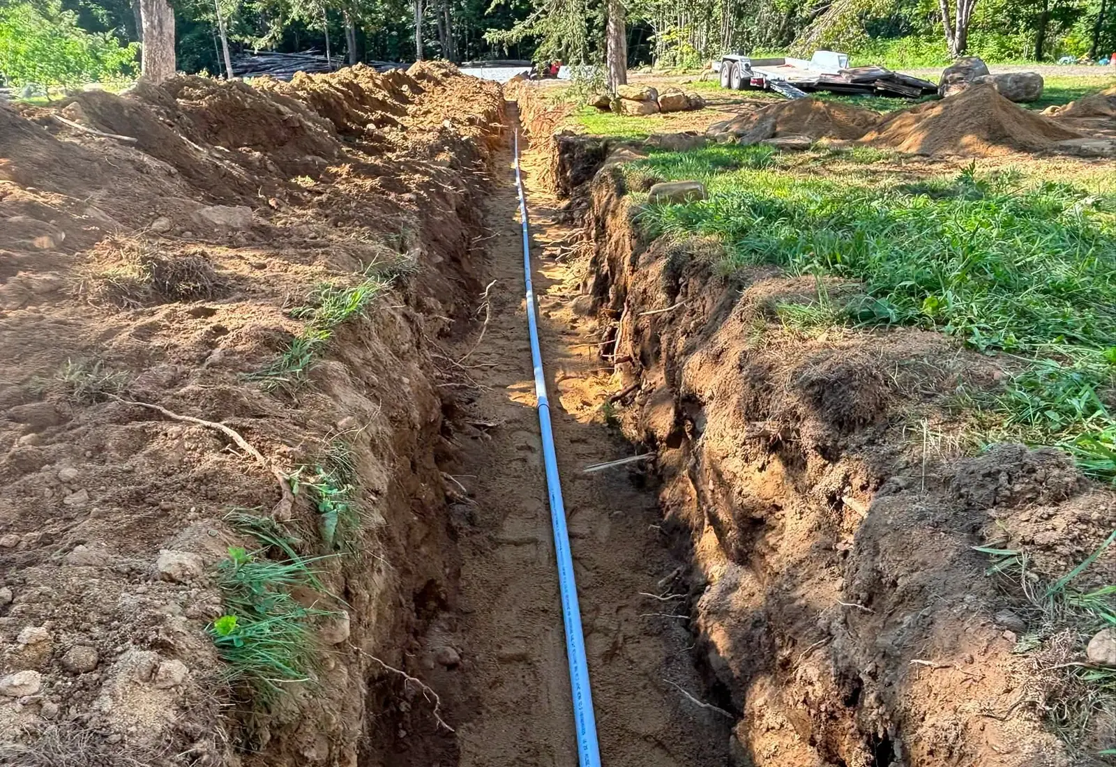 Grey electrical conduit running along the bottom of an open trench dug across a lawn, spoil piled on both sides